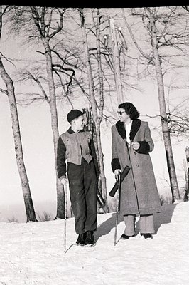 A man and woman, dressed in winter attire, stand on a snowy landscape. The man wears a dark suit and cap; the woman a long co...