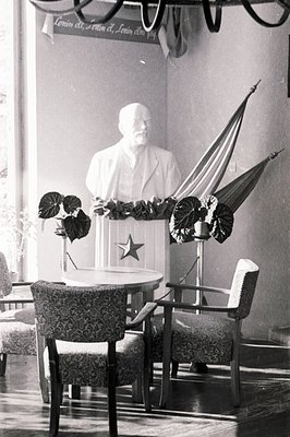 A bust of Lenin stands on a table adorned with a star, within a formally decorated room. Two dark chairs flank the table. A b...