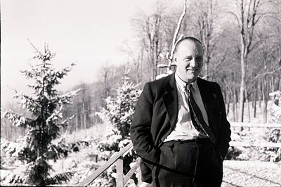 A formally dressed man stands outdoors, leaning against a snow-dusted evergreen. He wears a suit, tie, and appears relaxed. A...