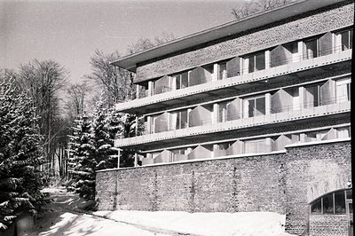A three-story brutalist building with projecting balconies dominates the view. Brick facade meets a snow-covered landscape an...