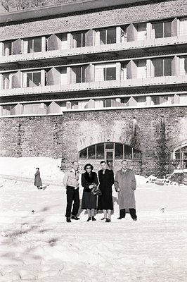 Four figures stand formally before a mid-century modern brick building with tiered balconies, likely a hotel or apartment com...