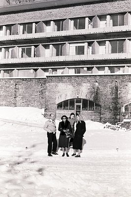 Three formally dressed individuals stand before a modernist brick building with projecting balconies. Heavy snowfall blankets...