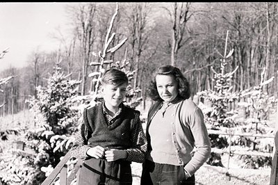 Two figures, a young boy and a young woman, stand in a snowy, wooded landscape. The boy wears a vest and checkered shirt, the...