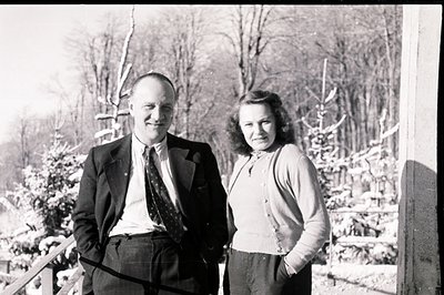 A formally-dressed man and woman stand outdoors, likely in a snowy, forested region. Man wears a suit, tie, and collared shir...
