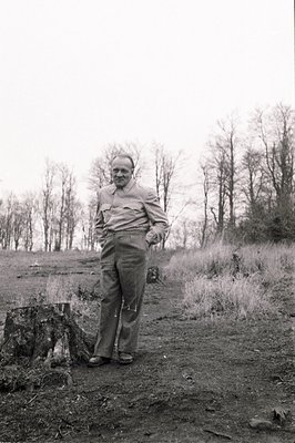 A man in workwear stands near a tree stump in a clearing. He wears a collared shirt, high-waisted trousers, and sturdy shoes....