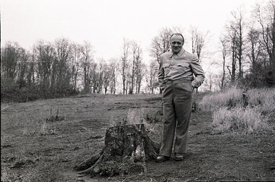 A man in workwear stands near a tree stump in a clearing, looking directly at the camera. High-waisted trousers, button-down ...