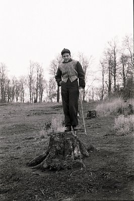 A young man stands on a tree stump, holding a walking stick. He wears a dark beret, a patterned vest over a collared shirt, a...