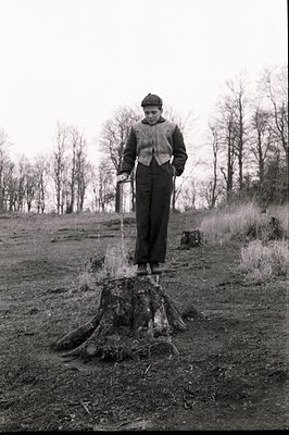 A young boy stands atop a weathered tree stump, balancing with apparent ease. He wears a tweed jacket, dark trousers, and a c...