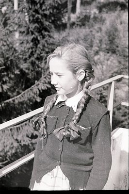 A young girl stands in profile, braided hair adorned with a large bow. She wears a dark blazer over a collared shirt and plai...