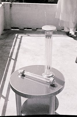 A mid-century modern outdoor scene shows a table with a tall glass sculpture and a low-slung glass vase. The architecture sug...