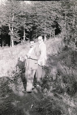 Two formally dressed men stand on a grassy slope overlooking a dense, evergreen forest. Both wear suits and eyeglasses; one h...