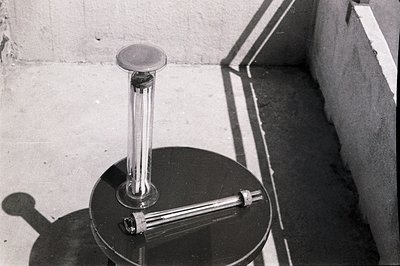 A black circular pedestal supports a tall, cylindrical form topped with a rounded cap. A shorter, horizontal cylinder extends...