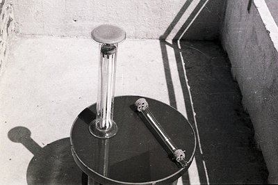 Black and white image showcasing scientific glassware on a circular lab bench. Two long test tubes are visible, one upright w...