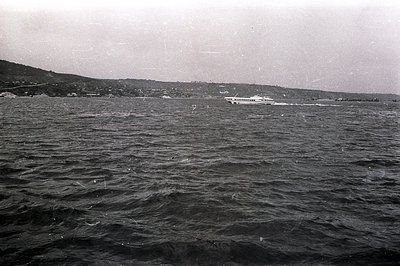 A black and white photograph captures a high-speed hydrofoil ferry traversing a choppy body of water. Distant coastline and b...