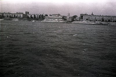 A black and white image captures a harbor scene, likely from the 1960s or 70s. A large passenger ship is docked alongside a w...