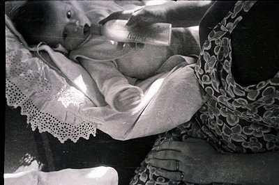 Infant being bottle-fed, cradled in a patterned shawl. Delicate lace trim adorns the baby's garment. The image exhibits chara...