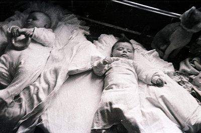 A monochrome image depicts a baby lying in a crib, swaddled in white fabric. Detail shows fine lace trim on the bedding and g...
