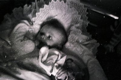 Vintage black and white portrait of a baby swaddled in a lace-trimmed blanket. Likely a formal studio portrait, evidenced by ...