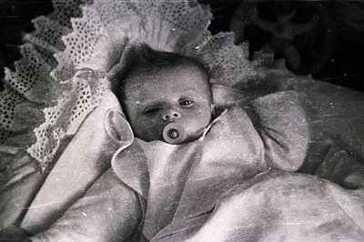 A monochrome portrait of an infant lying on a lacy surface, holding a pacifier. The image exhibits signs of age, including gr...
