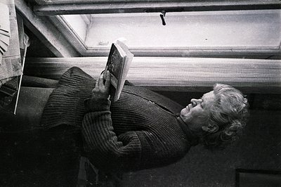 A man reclines, reading a book with evident concentration. He wears a thick, cable-knit sweater and rests on stacked newspape...