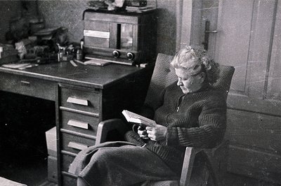 A seated woman with curly hair reads a document in a wood-framed rocking chair. A desk with drawers and an old radio are visi...