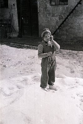 A young child, bundled in a knit sweater and hat, stands in a snowy courtyard. The child appears to be shielding their face w...