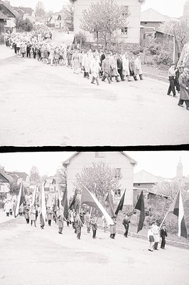 A long procession of individuals marches along a paved road, carrying flags with varying designs. Buildings line the street, ...