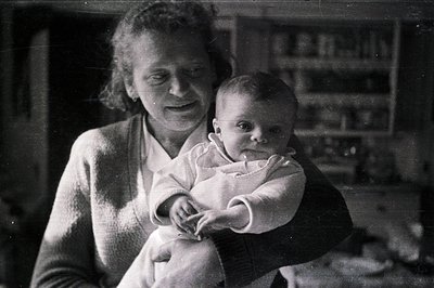 A young woman cradling an infant in a warmly lit interior. The woman wears a cable-knit sweater, and the baby is dressed in a...