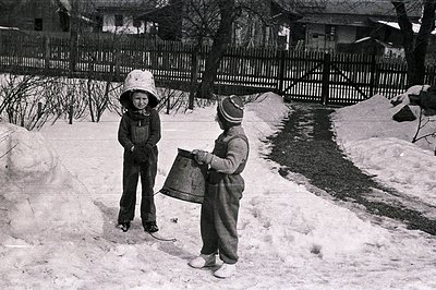 Two children in winter clothing stand in a snow-covered yard, engaged in play. One wears a large, hooded bucket; the other ca...