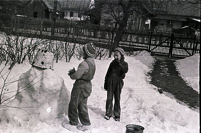 Two children, bundled in winter clothing, stand before a newly-built snowman in a snowy yard. A weathered wooden fence and a ...