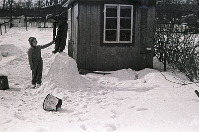 A boy stands on a large snow mound, reaching towards a small, dark-wooded building with a single window. He wears a knit hat ...
