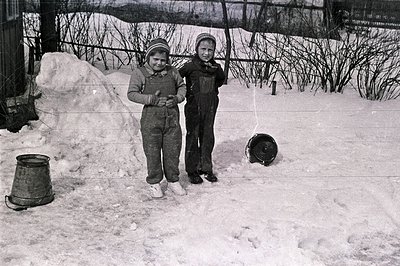 Two young children, bundled in winter clothing, stand in a snow-covered yard. A large snowdrift dominates the background, wit...