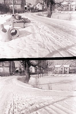 A snow-covered rural street scene. Two small houses flank a central road, lined with leafless trees and a large, prominent tr...