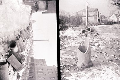 Black and white image depicts a snowy, rural landscape, likely post-construction. Numerous large concrete pipes are stacked a...
