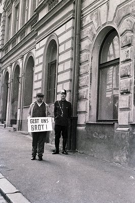 A man holding a sign reading "Gebt uns Brot!" (Give us bread!) stands alongside a uniformed police officer. The scene is set ...