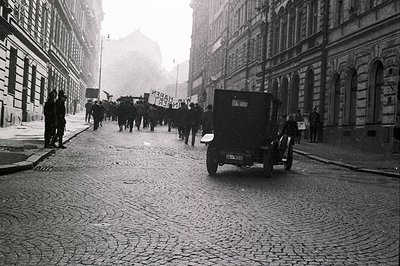 A formal procession moves along a cobblestone street lined with tall, ornate buildings. An open-top car with license plate "I...