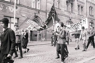 A black and white photograph captures a protest march with demonstrators carrying a banner reading "Geist - Brot - Frieden" (...