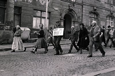 A group of people march along a cobblestone street, carrying a sign reading "Gebt uns Brot!" (Give us bread!). The architectu...