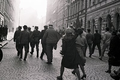 Procession of men and a young girl march along a cobblestone street, framed by neoclassical architecture with arched windows....