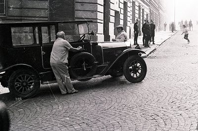 A man assists a driver with a vintage automobile, possibly a 1930s Bentley, on a cobbled street lined with buildings. Several...