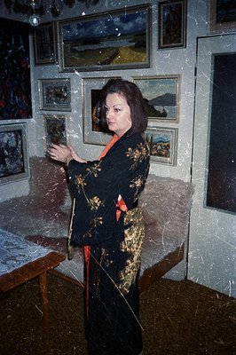 A woman in a richly embroidered black kimono stands in a room densely filled with framed landscape paintings. The room appear...