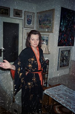 A woman in a patterned, dark-colored robe with a red lining stands within a room adorned with multiple framed artworks. The d...