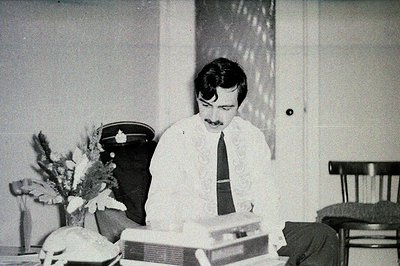 Man in suit sits at a typewriter, appearing deep in thought. An ornate floral arrangement and a rotary phone add vintage char...