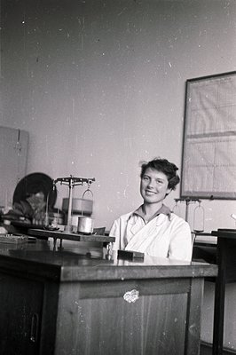 Young woman in lab coat, smiling, stands behind a workbench. Visible equipment includes a balance scale, suggesting a scienti...