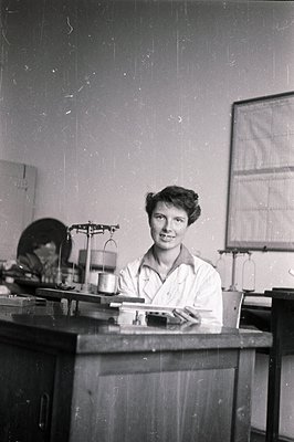 A young woman in a lab coat smiles while seated at a lab bench with scientific equipment. Likely a researcher or lab assistan...