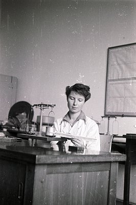 A young woman in a lab coat appears to be weighing a substance on a balance scale in a laboratory setting. The lab features p...