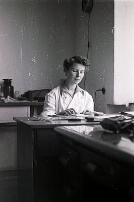 A young woman in a lab coat sits at a table strewn with papers, seemingly engrossed in her work. Likely a scientist or resear...