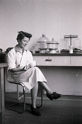 A seated woman in a lab coat contemplates a small vial; evident lab equipment and glassware behind. Likely a 1950s or 60s sci...