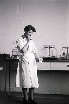 Woman in lab coat examines a flask within a lab setting. Features visible equipment like condensers & lab benches. Appears to...