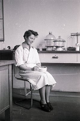 A seated woman in a lab coat examines a petri dish, likely conducting research. Minimalist chrome chair and utilitarian lab e...